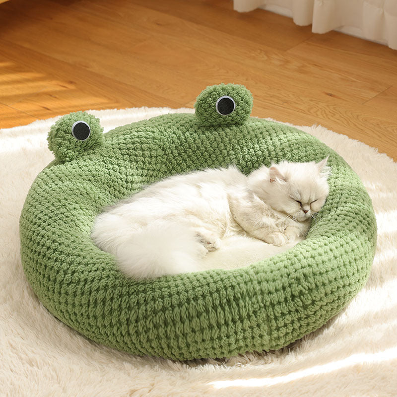 White cat lying on a green frog-shaped pet bed on a wooden floor.